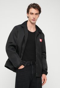 Μαύρο ελαφρύ μπουφάν, κλείσιμο με κουμπιά τύπου snap, μυτερός γιακάς, ελαστικές μανσέτες, διακριτική γυαλάδα, με κόκκινο λογότυπο στο αριστερό στήθος.