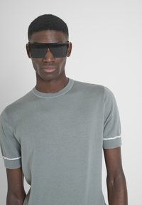 Maglietta grigia lavorata a maglia con maniche corte e colletto rotondo; presenta strisce bianche sui bordi dei polsini. Gli occhiali da sole neri angolari completano il look.