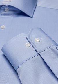 Helles blaues Hemd mit strukturiertem Kragen, zwei Knopfmanschetten, glatter Stofftextur und subtilem diagonalen Muster.