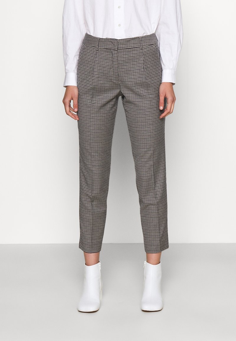 Femme portant un pantalon à carreaux slim longueur cheville avec une chemise blanche boutonnée et des bottines blanches, debout devant un fond uni.