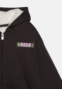 Tmavohnedá mikina na zips so bielou chlpaticou vnútri a vyšitým logom Roxy v ružovej a zelenej farbe na hrudi. Obsahuje predné vrecká.