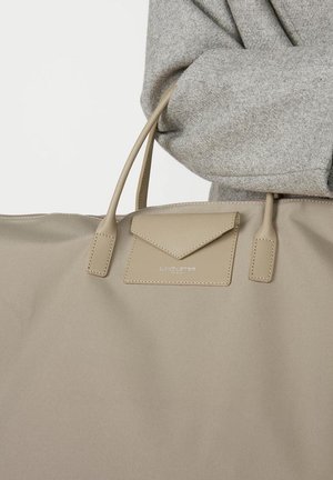 Sac fourre-tout beige en tissu lisse, avec deux poignées, une poche à rabat avec un design triangulaire et un logo en relief.