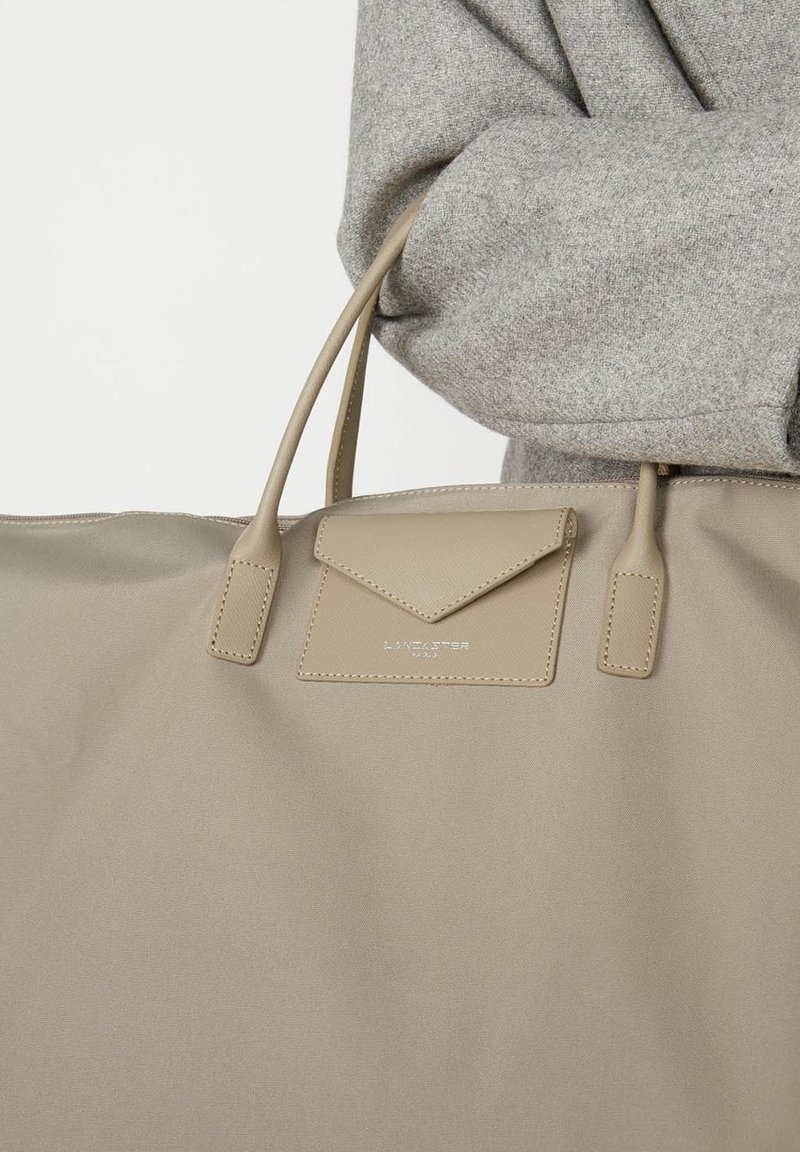 Sac fourre-tout beige en tissu lisse, avec deux poignées, une poche à rabat avec un design triangulaire et un logo en relief.