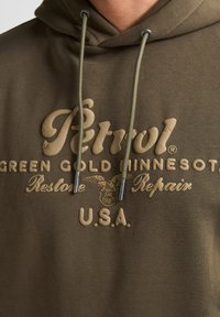 Olivově zelená mikina s klokaní kapsou, s béžovým embosovaným nápisem 'Petrol Green Gold Minnesota' a táhly na šňůrkách s kovovými akcenty.