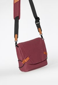 Bordeaux messenger taske med en glat overflade, brune læderdetaljer og en justerbar sort rem med metalbeslag og præget logo.