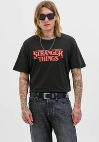 Černé bavlněné tričko s červeným nápisem "STRANGER THINGS", v kombinaci s tmavým džínovým kalhotami a černým pásem se stříbrnou přezkou; sluneční brýle nasazené.