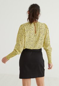 Blusa gialla con pois neri, maniche lunghe e spalle plissettate, abbinata a una gonna nera con dettaglio a chiave sulla schiena.