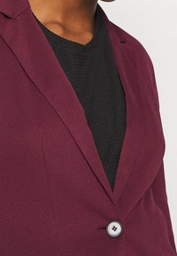 Burgundy texturerad kavaj med spetsiga slag, med en rund knappstängning och svart stickad innertopp. Nära vy av tygdetaljer.