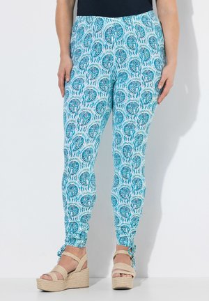 Vrouw draagt lichtblauwe legging met turquoise spiraalschelppatronen en beige sleehakken met enkelbandjes.