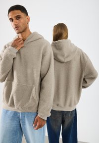 Beige flīsa pulovers ar kapuci, liels priekšējais kabatas, aukliņas kapucis un ribbas formas aproces. Modelis apvienojis ar gaiši ziliem un tumši ziliem džinsiem.