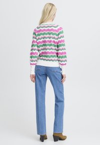 Pull multicolore avec un motif en zigzag rose, vert et blanc, assorti à un pantalon en jean bleu clair et des bottes marron à talons.