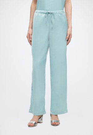 Žena oblečená v svetlomodrých širokých nohaviciach so sťahovaním a strieborných sandáloch na širokom opätku s otvorenou špičkou na bielom pozadí.