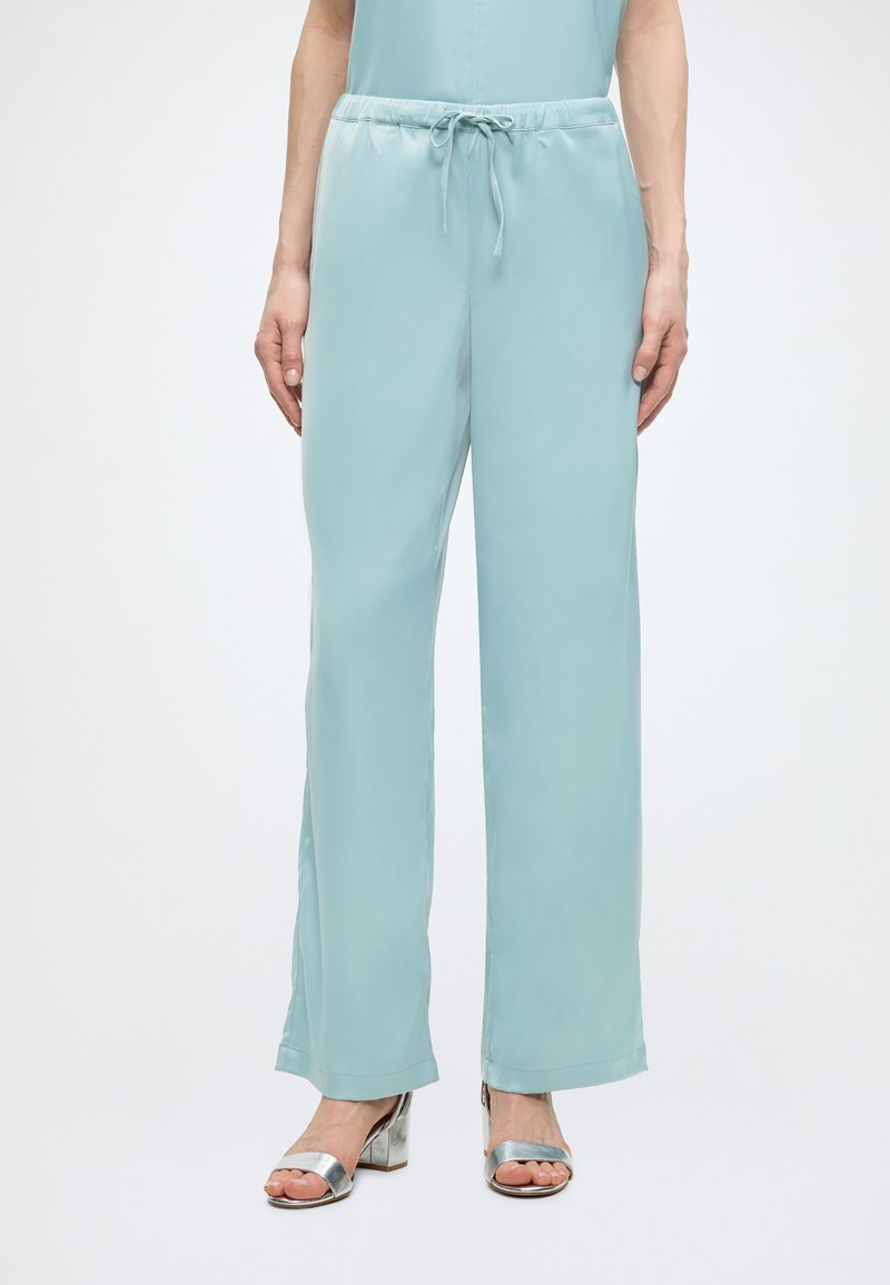 Femme portant un pantalon large bleu clair à cordon et des sandales argentées à talon bloc et orteils ouverts sur fond blanc.