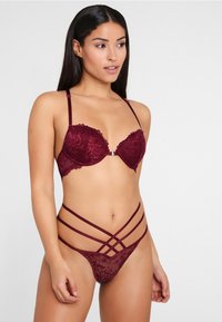 Bordeaux Spitzen-BH mit Unterdraht und verstellbaren Trägern; passender Tanga mit mehreren Übere Kreuzgurtungen und Spitzenverzierungen.