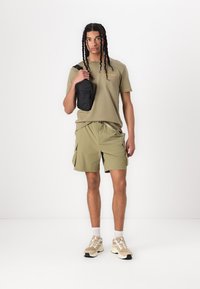 Olijfgroen t-shirt met klein logo, bijpassende cargo shorts en beige sportschoenen. Zwarte schoudertas; schoon, minimalistisch ontwerp.