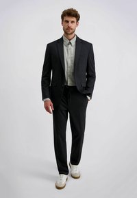 Man loopt in een donker pak met een lichtgrijze blouse en witte sneakers voor een effen lichte achtergrond.
