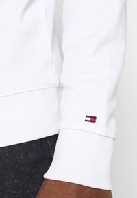 Felpa bianca con polsini a coste, caratterizzata da un piccolo logo ricamato in rosso, bianco e blu sulla manica sinistra. Texture morbida.