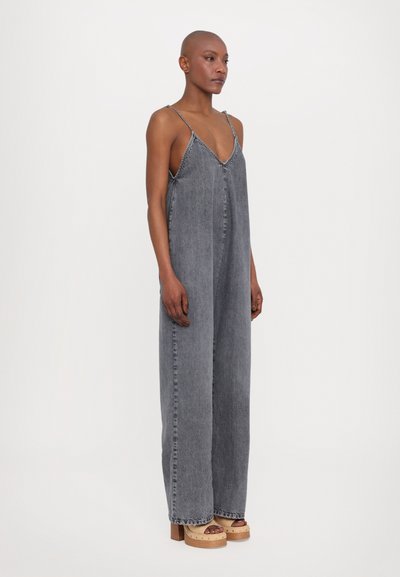 Combinaison en denim gris avec un décolleté en V profond, des bretelles spaghetti et des jambes larges. Présente des coutures visibles et une coupe décontractée. Associée à des sandales à plateforme en bois.