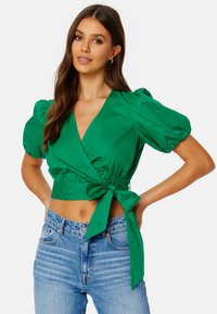 Top avvolgente verde con maniche a sbuffo corte, scollatura a V profonda e dettaglio in vita annodato. Abbinato a jeans blu sbiaditi a vita alta.