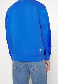 Blå sweatshirt med en strukturerad, mjuk tygkvalitet. Har kontrasterande glänsande blå ärmar och en vit logodetalj nära fållen. Lätt och ledig design.