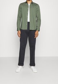 Man in olijfgroene button-up overhemd over een wit T-shirt, zwarte broek en witte sneakers, staande tegen een effen achtergrond.