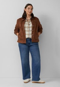 Bruine suède jas met een kraag, knoopsluiting en twee voorzakken, gedragen over een geruite blouse en een blauwe wijde broek. Witte sneakers.