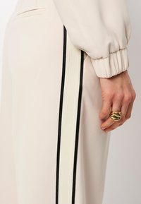 Beige broek met zwarte en crèmekleurige zijstrepen. Zachte, gladde textuur. Hand versierd met een chunky gouden ring.