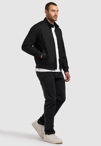 Mees kõnnib, seljas must bomber-jakk valge t-särgi, mustade teksade ja valgete tennistega heledal hallil taustal.