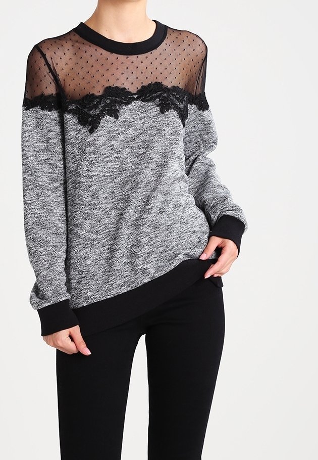 Grijze sweatstof met textuur, voorzien van een zwarte kanten rand en een doorschijnend zwart mesh paneel bovenop. Lange mouwen met zwarte manchetten, gecombineerd met zwarte broek.