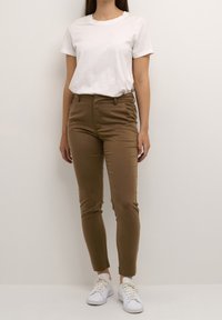 Brown fitted pants made of stretch fabric, featuring a mid-rise waist and side pockets, paired with a plain white t-shirt and white sneakers.