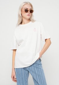 Hvid bomulds T-shirt med korte ærmer og et lille pink motiv på venstre bryst. Matchet med lyseblå jeans med lodrette striber.