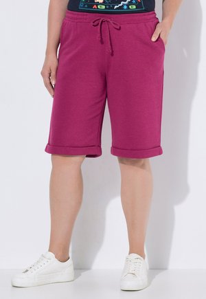 Osoba ubrana w magentowe szorty z podwiniętymi nogawkami i sznurkiem w talii oraz białe trampki, stojąca na jednolitym białym tle.