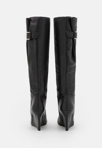 Par de botas altas de cuña de cuero negro con correas de hebilla lateral, mostradas desde atrás sobre un fondo blanco.