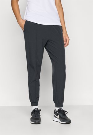 Pantalon de jogging gris avec poignets élastiques et poches latérales, fabriqué dans un tissu lisse. Associé à des baskets noires avec un accent blanc.