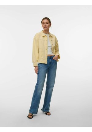 Femme debout portant une veste boutonnée jaune clair, un top court blanc, un jean bleu et des sandales noires à bout ouvert sur un fond blanc uni.