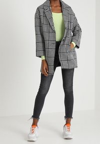 Cappotto oversize a scacchi bianco e nero, top verde, jeans scuri con orlo sfilacciato e sneakers bianche con dettagli arancioni.