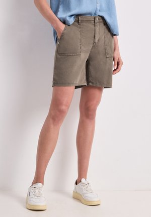 Person iført grønne knælange shorts med forlommer, en lyseblå skjorte og hvide sneakers, stående op ad en flad væg.