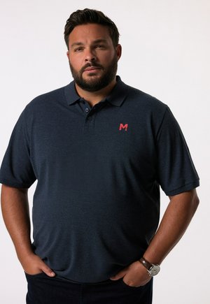 Polo blu navy con finitura testurizzata e maniche corte. Presenta un piccolo logo rosso sul petto. Indossato con jeans scuri e un orologio.