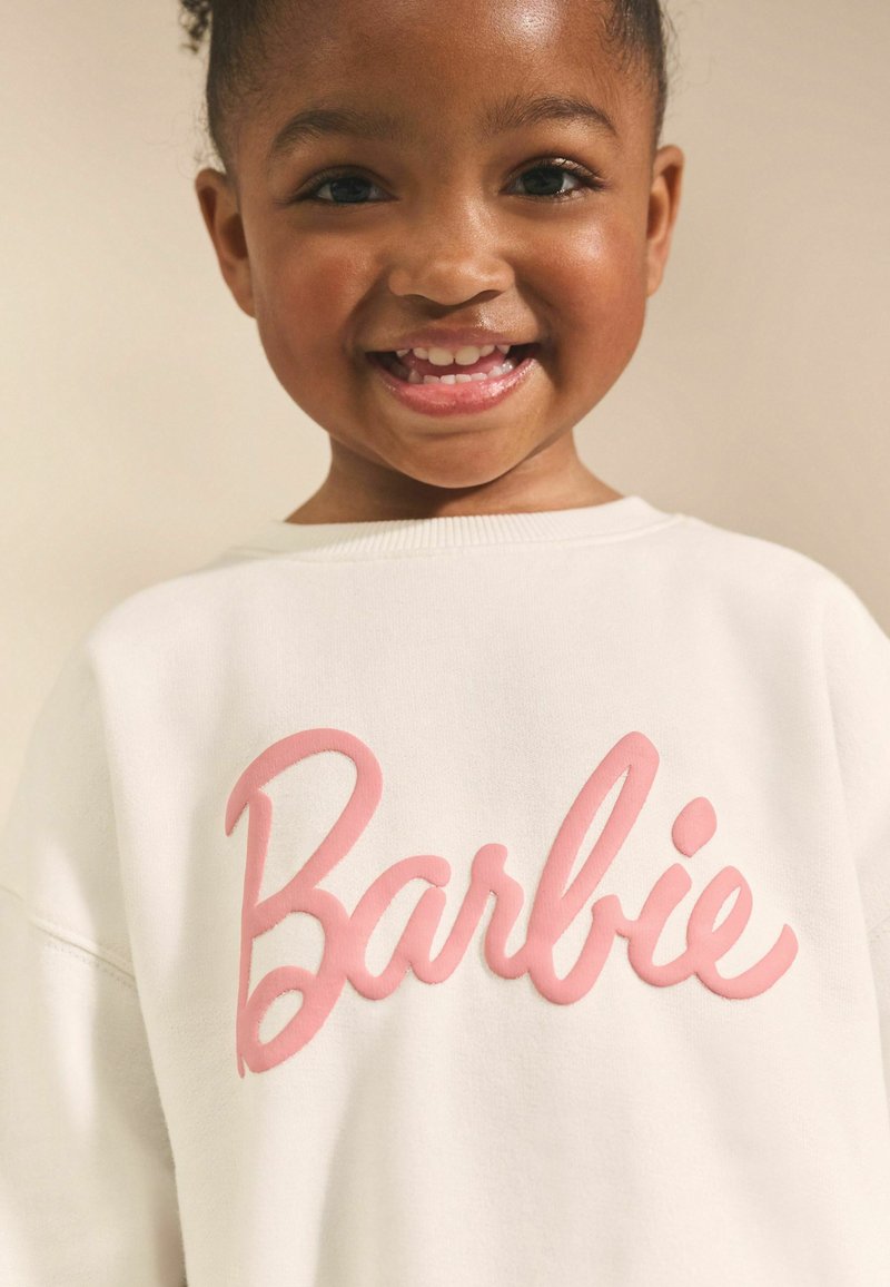 Bela majica s kapuljačom na kojoj se u debelim, ružičastim, izbočenim slovima nalazi reč "Barbie". Tkanina deluje mekano, s opuštenim krojem i okruglim izrezom.
