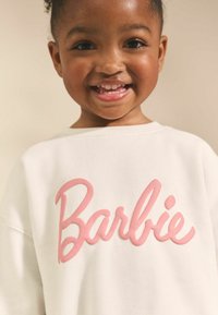 Hanorac alb cu cuvântul "Barbie" în litere îndrăznețe, roz, reliefate. Materialul pare moale, cu o croială lejeră și gât rotund.