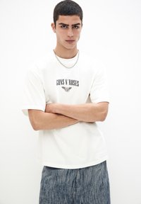 Hvid bomulds t-shirt med "GUNS N' ROSES" trykt i sort, parret med blå teksturerede shorts. Modellen bærer en sølv kæde halskæde.