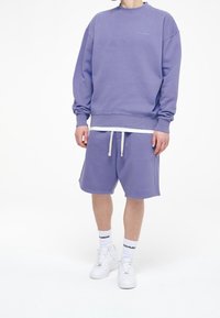 Uomo che indossa un felpa lilla e pantaloni corti abbinati, con sneakers bianche e calze bianche di marca su sfondo bianco.
