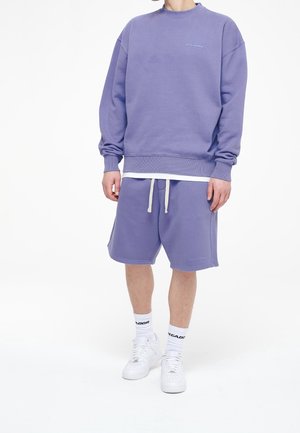 Mann trägt einen lilafarbenen Sweatshirt und passende Shorts mit weißen Sneakers und markierten weißen Socken auf weißem Hintergrund.