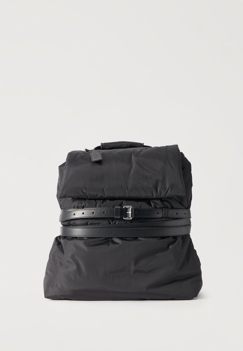 Μαύρο μαλακό σακίδιο με διπλωμένη κορυφή, στερεωμένο με δύο μαύρες δερμάτινες ζώνες με ασημένιες αγκράφες, σε απλό ανοιχτό φόντο.