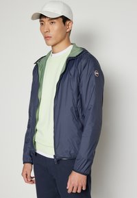 Marine lett jakke med grønn innvendig, glidelås foran, sidelommer og logo-patch på skulderen; brukt over en lysegrønn hettegenser.