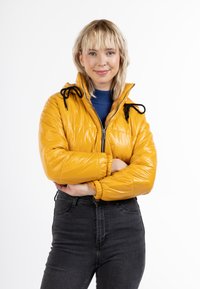 Gul puffjacka med en glänsande yta, justerbara svarta dragkedjor på huvan, dragkedjestängning och elastiska ärmslut. Bärs över en blå topp.