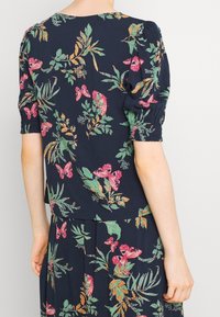 Blusa floreale blu navy con maniche a palloncino, decorata con farfalle rosa e motivi di fogliame verde. Realizzata in tessuto leggero, con texture e polsini elasticizzati.
