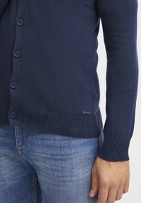 Mørkeblå cardigan laget av myk strikket stoff, med knappelukking på fronten, ribbede mansjetter og et merketap ved kanten. Sammen med jeans i denim.