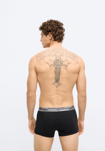 Fekete boxeralsó szürke elasztikus derékpánttal, logóval díszítve. Egy modell mezítelen hátán bemutatva, ahol egy nagy tetoválás látható.