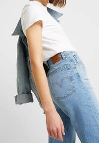 Woman wearing light blue high-waisted jeans, white t-shirt, and denim jacket draped over shoulders against a plain background.